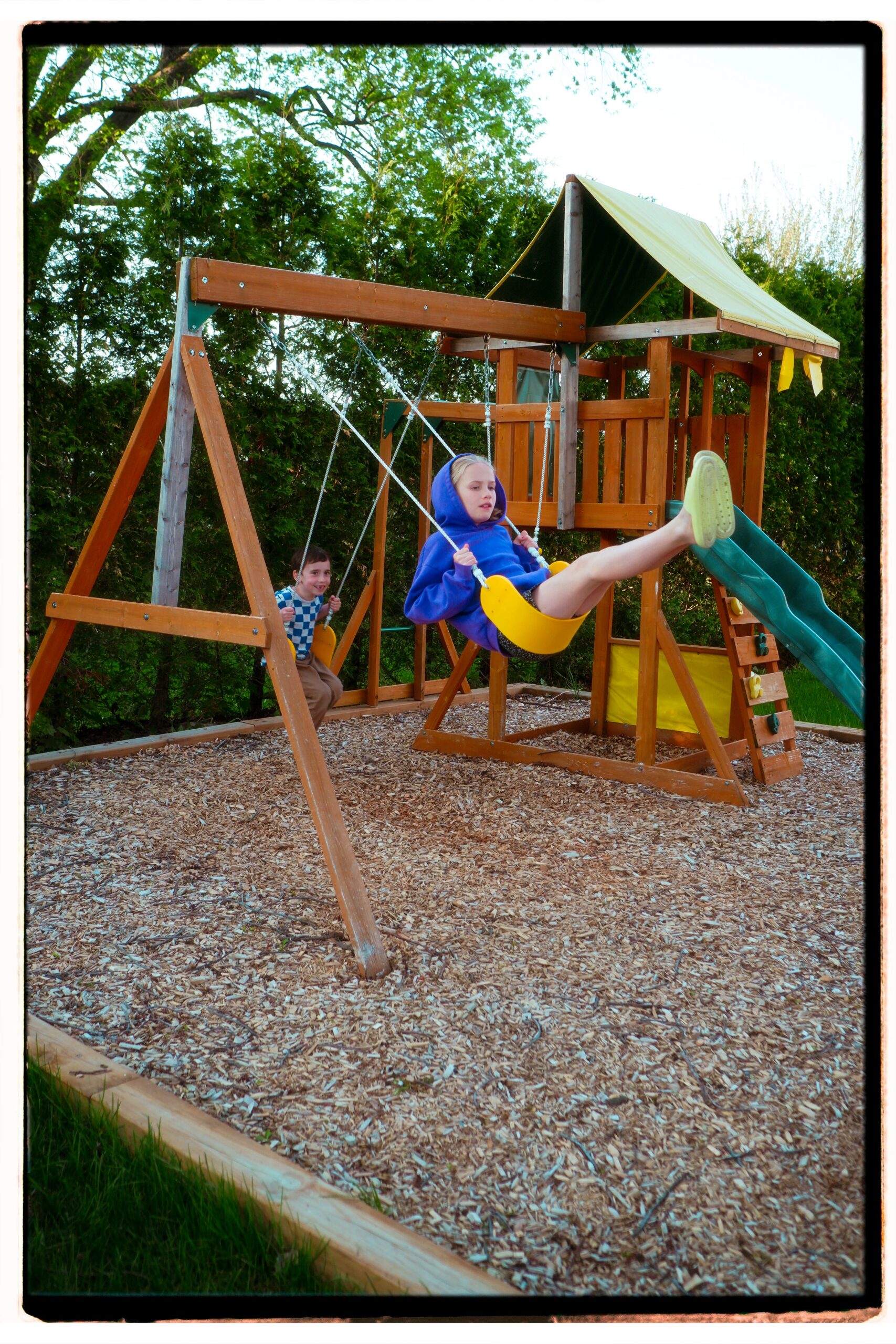 Two children playing on a backyard wooden swing set with a shade canopy